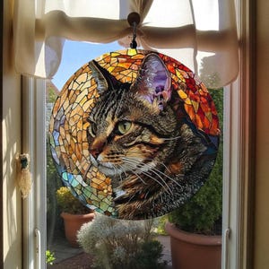 May include: A round stained glass window hanging featuring a detailed tabby cat portrait. The cat has green eyes and is set against a mosaic background of warm orange, yellow, and brown tones. The window is suspended in front of a window.