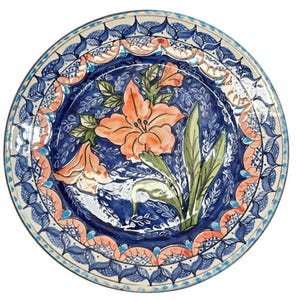 May include: Round ceramic plate with a hand-painted floral design. The plate features a vibrant orange flower with green leaves against a deep blue background. The rim is decorated with intricate blue and peach patterns, creating a decorative piece.