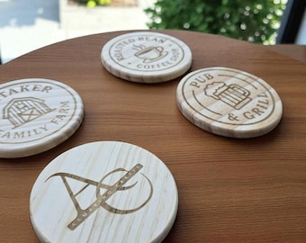 Könnte beinhalten: Vier runde Untersetzer aus Holz mit eingravierten Designs. Ein Untersetzer trägt die Aufschrift "Whitaker Family Farm" mit einer Scheunen-Grafik. Ein anderer trägt die Aufschrift "Roasted Bean Coffee Shop" mit einer Kaffeetasse. Der dritte trägt die Aufschrift "Pub & Grill" mit einem Bierkrug. Der letzte hat ein Monogramm "A" und "C".