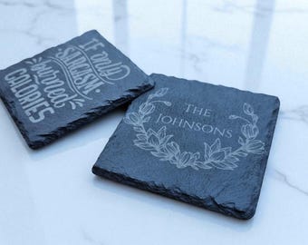 Puede incluir: Dos posavasos cuadrados de pizarra. Un posavasos tiene el texto "If only sarcasm burned calories" en escritura blanca. El otro posavasos tiene el texto "The Johnsons" dentro de un diseño de corona floral. Ambos son de color gris oscuro.