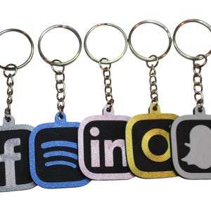 May include: Five keychains with silver-colored key rings and chains. Each keychain features a different social media logo: Facebook, Spotify, LinkedIn, Instagram, and Snapchat. The logos are in various colors, including silver, blue, pink, gold, and gray.
