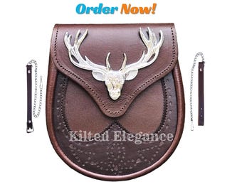 Könnte beinhalten: Eine braune Leder-Sporran mit einem silbernen Hirschkopf-Emblem. Die Sporran hat einen Klappenverschluss und dekorative Nähte. Der Text "Kilted Elegance" ist sichtbar. Enthält zwei Lederriemen mit Ketten.