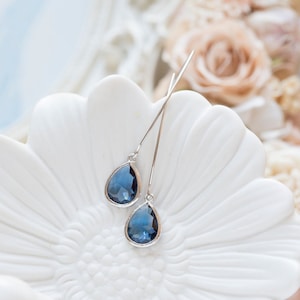 Puede incluir: Un par de pendientes de plata en forma de lágrima con detalles de cristal azul oscuro. Los pendientes se muestran sobre una superficie blanca en forma de flor.