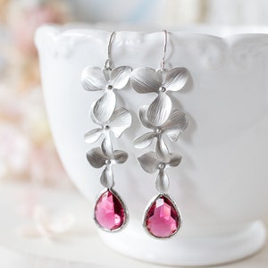 May include: Silver-coloured drop earrings featuring a floral design with flower-shaped elements and a magenta teardrop gemstone. The earrings are displayed in front of a white decorative bowl.