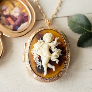 May include: Gold locket necklace with a cameo of a cherub in a floral design. The locket opens to reveal a space for a photo.