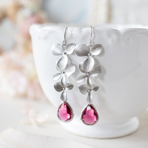 Może przedstawiać: Srebrne kolczyki z trzema delikatnymi wisiorkami w kształcie kwiatów i różową kropelką kryształu.