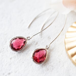 May include: A pair of drop earrings featuring faceted, teardrop-shaped deep pink stones set in silver-toned metal frames. The earrings hang from curved, slender silver-toned ear wires that extend upward, creating a long, elegant profile for the jewelry.