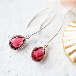 Può includere: Un paio di orecchini pendenti in argento con pietre preziose rosso scuro a forma di goccia. Gli orecchini hanno un design delicato, simile a un filo, e sono posti su uno sfondo bianco con un elemento decorativo dorato.