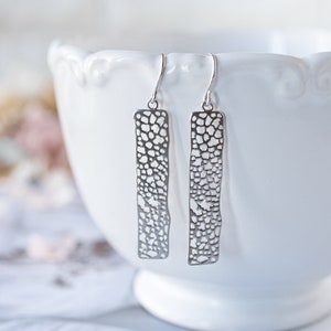 May include: A pair of silver dangle earrings with a delicate, openwork design. The earrings are rectangular in shape and feature a textured, patterned surface.