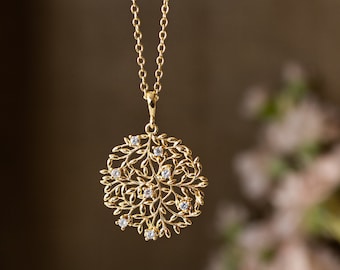 May include: A gold-colored pendant necklace featuring a circular, openwork design of leaves and small gemstones. The pendant hangs from a delicate chain. The background is a blurred, neutral color.