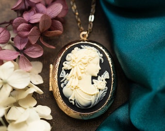 Puede incluir: Un collar de cadena dorada con un medallón de camafeo blanco y negro que presenta el perfil de una mujer. El medallón está rodeado por un marco dorado y tiene una bisagra en el lateral. El collar está sobre una superficie marrón con flores rosas y blancas.