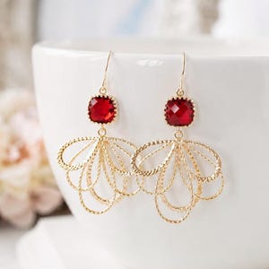 May include: A pair of gold-tone drop earrings featuring a square-cut red gemstone at the top, connected to a dangling, multi-layered fan-shaped charm made of thin, textured gold wire loops that create an open, airy, and intricate design.
