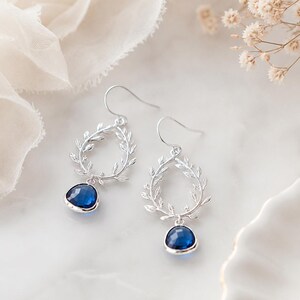May include: A pair of silver-toned drop earrings. Each earring features a teardrop-shaped frame adorned with leaf details, with a vibrant blue gemstone suspended at the bottom. The earrings are displayed on a white surface.
