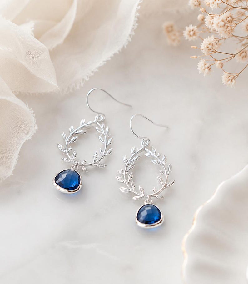 May include: A pair of silver-toned drop earrings. Each earring features a teardrop-shaped frame adorned with leaf details, with a vibrant blue gemstone suspended at the bottom. The earrings are displayed on a white surface.