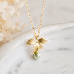 May include: A gold-coloured necklace with a floral pendant and a light green pearl. The necklace is displayed on a white marble surface. The pendant has three petals. Dried flowers are in the background.