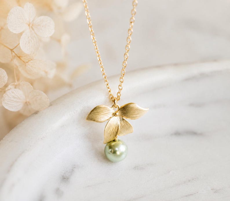 May include: A gold-coloured necklace with a floral pendant and a light green pearl. The necklace is displayed on a white marble surface. The pendant has three petals. Dried flowers are in the background.