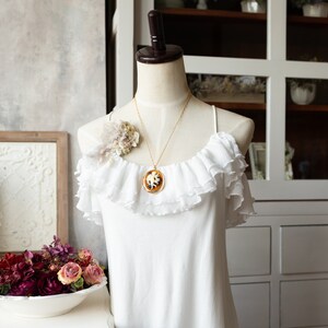 May include: A white off-the-shoulder top with ruffles and a gold chain necklace with a cameo pendant. The necklace features a white cameo with a brown design.