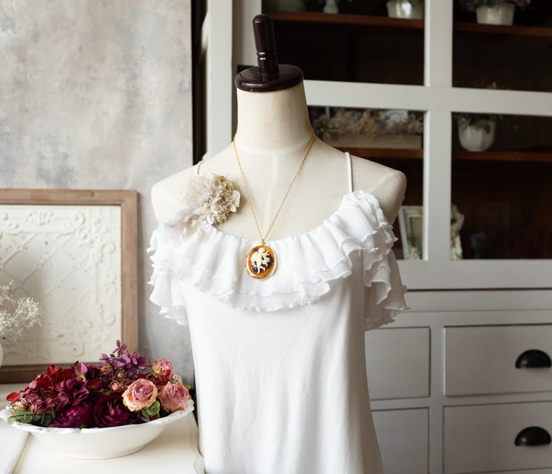 May include: A white off-the-shoulder top with ruffles and a gold chain necklace with a cameo pendant. The necklace features a white cameo with a brown design.