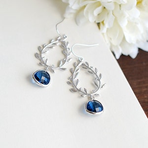 May include: Silver-toned dangle earrings with a leaf design and teardrop-shaped, faceted, dark blue gemstones. The earrings are displayed on a white surface with a blurred background of a book and white flowers.