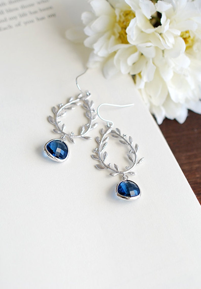 May include: Silver-toned dangle earrings with a leaf design and teardrop-shaped, faceted, dark blue gemstones. The earrings are displayed on a white surface with a blurred background of a book and white flowers.