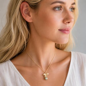 May include: A gold necklace with a pendant comprising three leaf shapes and a light green pearl. The necklace is worn by a person with blonde hair and a white top.