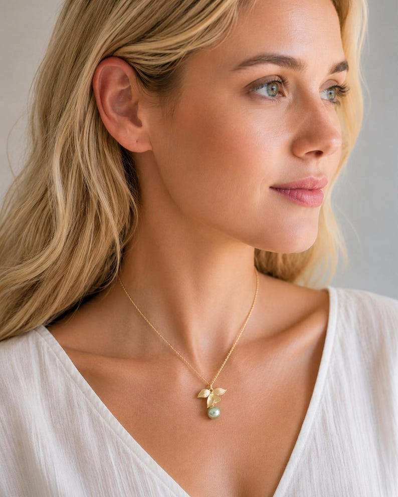 May include: A gold necklace with a pendant comprising three leaf shapes and a light green pearl. The necklace is worn by a person with blonde hair and a white top.