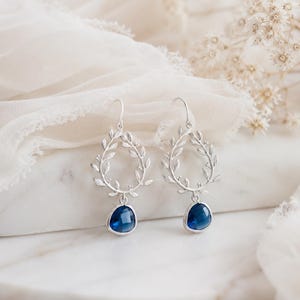 May include: Silver-toned drop earrings featuring a leaf-like wreath design. Each earring has a teardrop-shaped, deep blue gemstone. The earrings are displayed on a white marble surface with a soft, white fabric background.