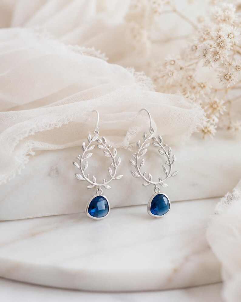 May include: Silver-toned drop earrings featuring a leaf-like wreath design. Each earring has a teardrop-shaped, deep blue gemstone. The earrings are displayed on a white marble surface with a soft, white fabric background.
