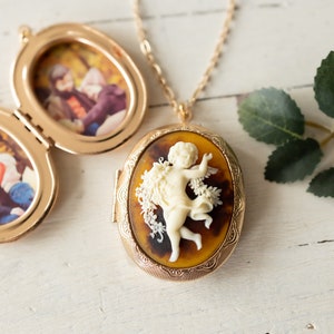 May include: A gold locket necklace with a cameo of a cherub. The locket opens to reveal a photo insert. The locket is on a white surface with green leaves.