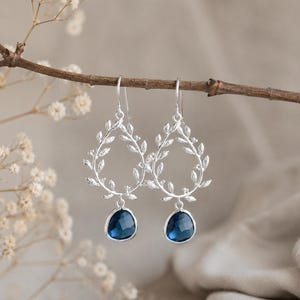 May include: Silver-toned earrings with a leaf design and teardrop-shaped, dark blue gemstones. The earrings are suspended from a branch with small white flowers in the background. The earrings are a delicate and elegant accessory.