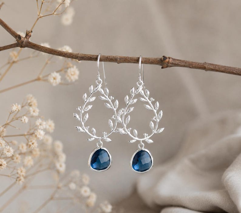 May include: Silver-toned earrings with a leaf design and teardrop-shaped, dark blue gemstones. The earrings are suspended from a branch with small white flowers in the background. The earrings are a delicate and elegant accessory.