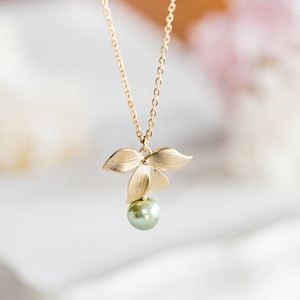 May include: A gold-tone necklace with a pendant shaped like three leaves, holding a single, round, pale green pearl. The chain is fine, and the pendant has a matte finish. The necklace is set against a soft, blurred background.