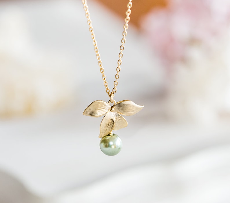 May include: A gold-tone necklace with a pendant shaped like three leaves, holding a single, round, pale green pearl. The chain is fine, and the pendant has a matte finish. The necklace is set against a soft, blurred background.