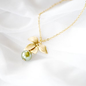 May include: A gold necklace with a floral pendant and a green pearl. The pendant has three textured leaves and a small, round pearl. The necklace is shown against a white, silky background.
