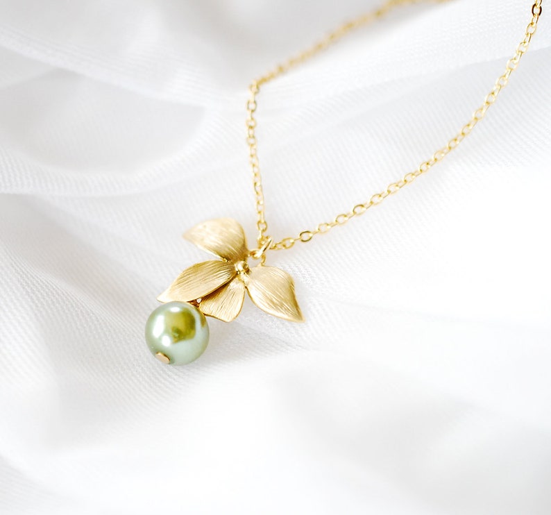 May include: A gold necklace with a floral pendant and a green pearl. The pendant has three textured leaves and a small, round pearl. The necklace is shown against a white, silky background.