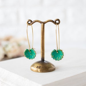 Op de afbeelding: Een paar gouden oorhangers met groene, bladvormige bedels. De oorhangers worden getoond op een gouden standaard.