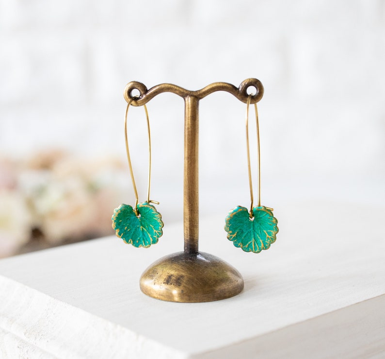 Op de afbeelding: Een paar gouden oorhangers met groene, bladvormige bedels. De oorhangers worden getoond op een gouden standaard.