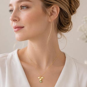 May include: A gold necklace featuring a leaf-shaped pendant with a small green gemstone. The pendant has three gold leaves and a small green stone. The necklace is worn by a person wearing a white top.