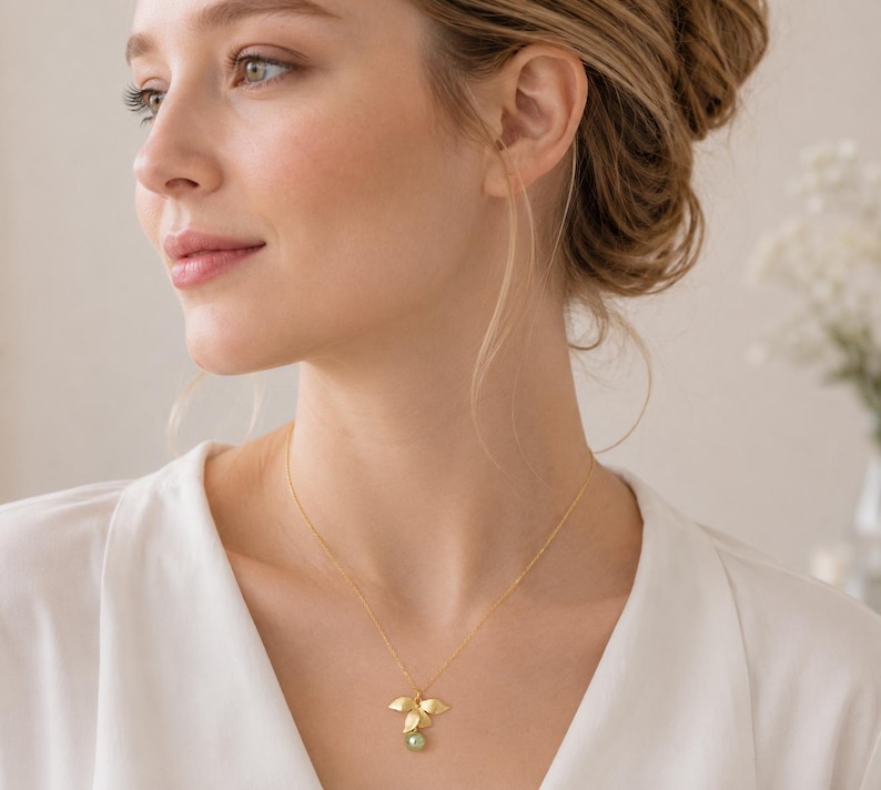 May include: A gold necklace featuring a leaf-shaped pendant with a small green gemstone. The pendant has three gold leaves and a small green stone. The necklace is worn by a person wearing a white top.