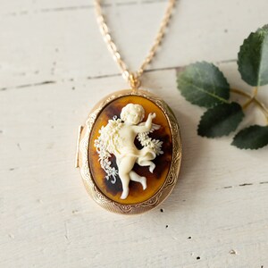 May include: A gold chain necklace with a cameo locket featuring a white cherub figurine with wings and a floral design. The locket has a brown and gold patterned background.
