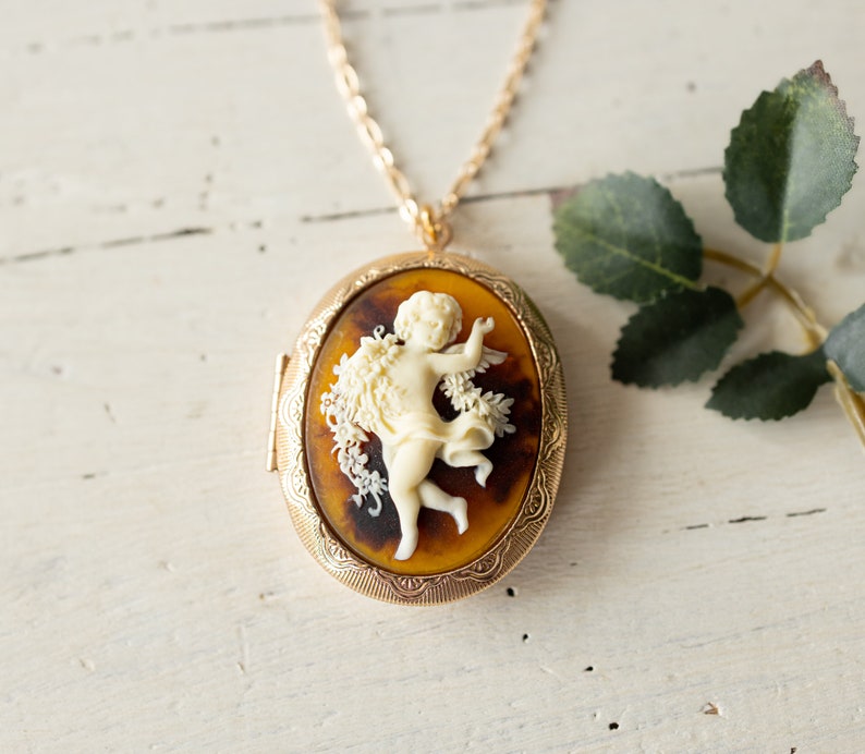 May include: A gold chain necklace with a cameo locket featuring a white cherub figurine with wings and a floral design. The locket has a brown and gold patterned background.