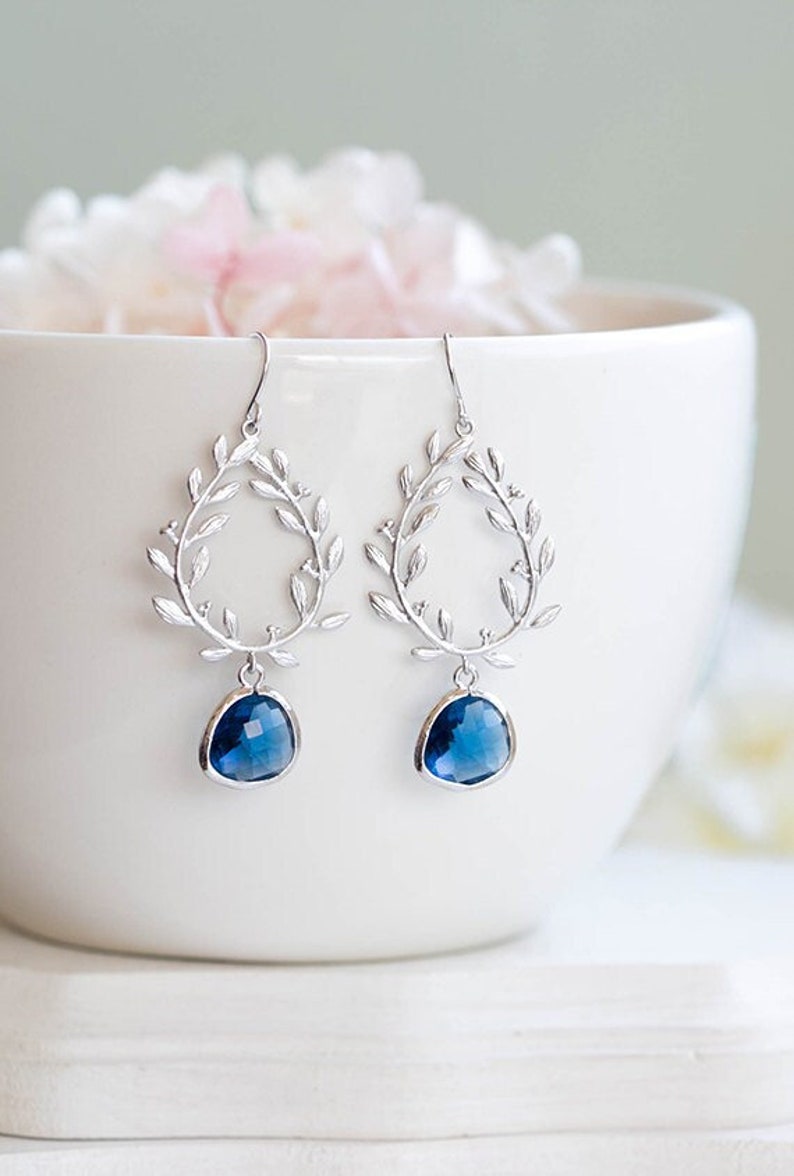 May include: Silver-toned drop earrings with a leaf-like design. Each earring features a teardrop-shaped, faceted, deep blue gemstone. The earrings are displayed against a white background, with soft pink and white flowers in the background.