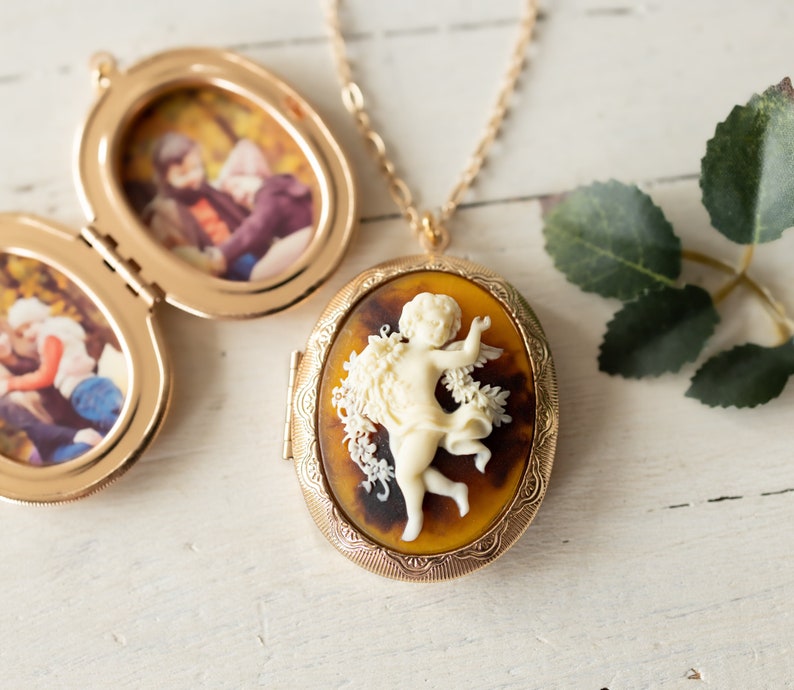 May include: Gold locket necklace with a cameo of a cherub in a floral design. The locket opens to reveal a space for a photo.