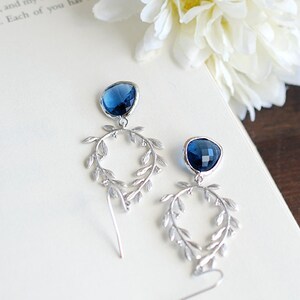 May include: A pair of silver-toned drop earrings with a dark blue gemstone at the top and a leaf-like design. The earrings are on a white surface with a flower and text in the background. The text reads "with distress..." and "Each of you has my".
