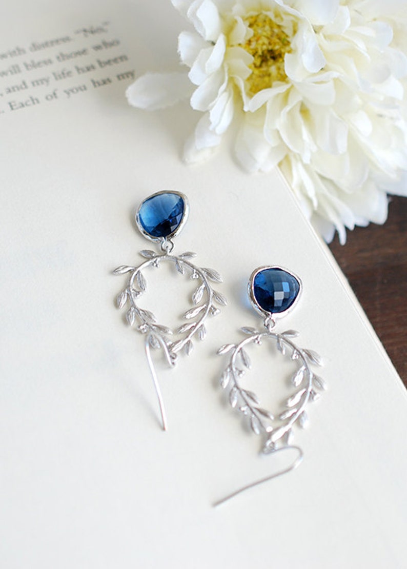 May include: A pair of silver-toned drop earrings with a dark blue gemstone at the top and a leaf-like design. The earrings are on a white surface with a flower and text in the background. The text reads "with distress..." and "Each of you has my".