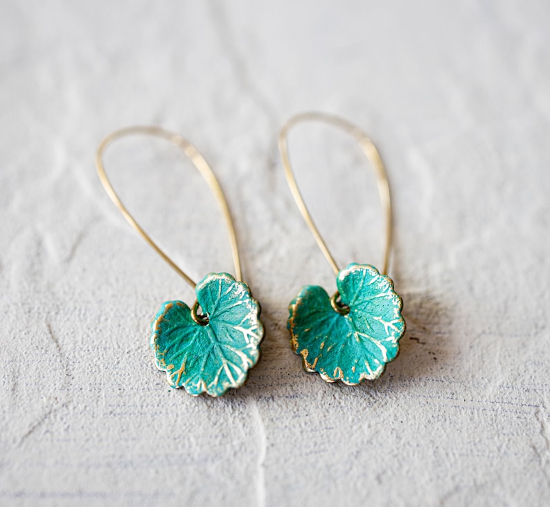 Op de afbeelding: Een paar gouden oorbellen met turquoise groene bladeren als hanger. De bladeren hebben een getextureerd oppervlak en een gouden rand.