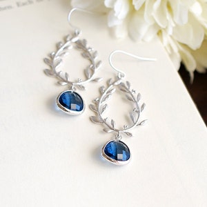 May include: Silver-toned earrings featuring a laurel leaf design. Each earring has a teardrop-shaped, faceted, deep blue gemstone. The earrings are displayed on a white surface with a blurred floral background.