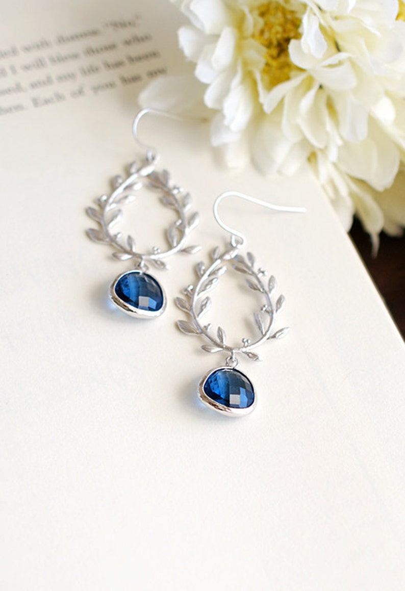 May include: Silver-toned earrings featuring a laurel leaf design. Each earring has a teardrop-shaped, faceted, deep blue gemstone. The earrings are displayed on a white surface with a blurred floral background.