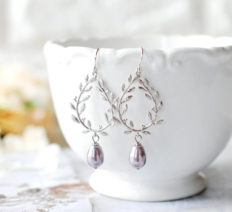 May include: A pair of silver-toned drop earrings. Each earring features a teardrop-shaped frame adorned with leaf details, suspending a lavender-colored pearlescent bead. The earrings are displayed against a blurred white background.
