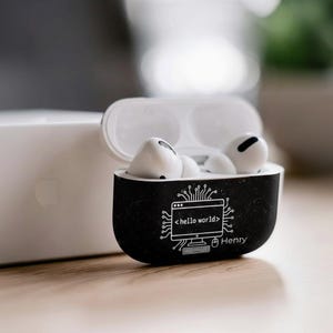 May include: White wireless earbuds in a black case with a white "hello world" computer graphic and the name "Henry". The case is open, showing the earbuds. A white Apple product is in the background.